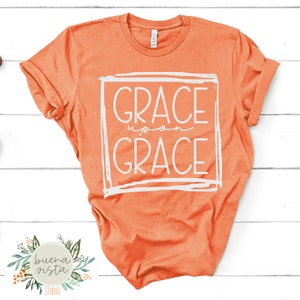 May include: Orange t-shirt with the words "GRACE upon GRACE" in a white, hand-drawn style within a white square. The shirt is displayed on a white wooden surface. The Buena Vista Studio logo is in the bottom left corner.