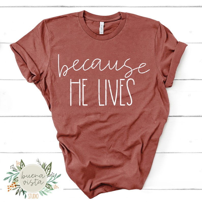 K&ouml;nnte beinhalten: Ein weinrotes T-Shirt mit dem wei&szlig;en Aufdruck "because HE LIVES". Das Shirt hat einen Rundhalsausschnitt und hochgekrempelte &Auml;rmel. Das Logo von Buena Vista Studio ist unten links zu sehen. Das T-Shirt ist aus weichem Stoff.