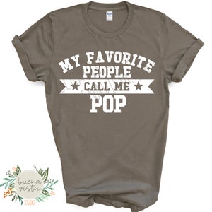 May include: A gray t-shirt with the text "MY FAVORITE PEOPLE CALL ME POP" in white block letters. The text is arranged in three lines with a banner and stars. The shirt has rolled-up sleeves.