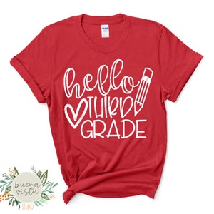 May include: A red t-shirt with the words "hello THIRD GRADE" in white script, a white heart, and a pencil illustration. The shirt is a crew neck style and has rolled-up sleeves. The Buena Vista Studio logo is in the bottom left corner.