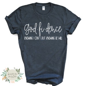May include: Dark blue t-shirt with the words "God-fidence" in a white script font. Below the main text is the phrase "Knowing I can't but knowing He will" in a smaller white font. The shirt has rolled-up sleeves.