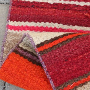 May include: A handwoven rug with a colorful striped pattern in shades of red, orange, brown, and white. The rug is folded in half, showing the intricate details of the weaving.
