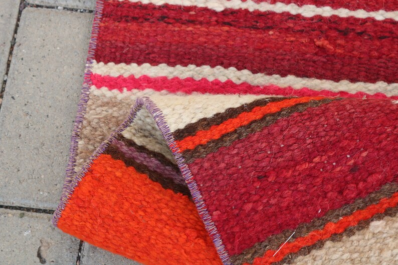 May include: A handwoven rug with a colorful striped pattern in shades of red, orange, brown, and white. The rug is folded in half, showing the intricate details of the weaving.
