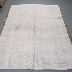 May include: A rectangular, off-white rug with a woven texture and a fringed border. The rug has a neutral color palette, suitable for various interior styles. The rug is likely made of natural fibers.