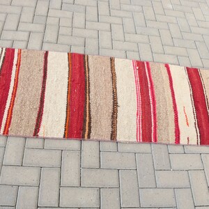 May include: A long, narrow, striped rug with red, brown, and white stripes. The rug is woven with a rough texture and has a fringe on the ends.
