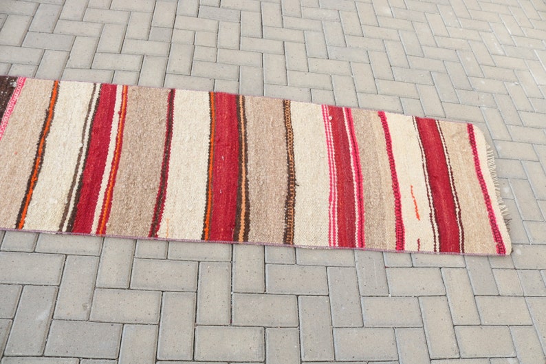 May include: A long, narrow, striped rug with red, brown, and white stripes. The rug is woven with a rough texture and has a fringe on the ends.