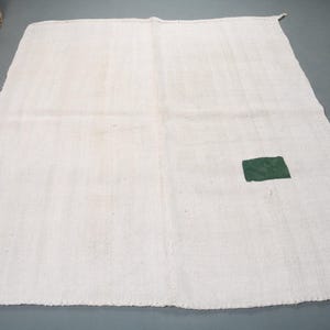 Może przedstawiać: Biały tkany dywan z małym kwadratowym zielonym łatką w środku.