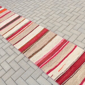 May include: A long, narrow, striped rug with a cream background and red, orange, and brown stripes. The rug is made of wool and has a fringe on both ends.
