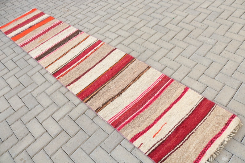 May include: A long, narrow, striped rug with a cream background and red, orange, and brown stripes. The rug is made of wool and has a fringe on both ends.