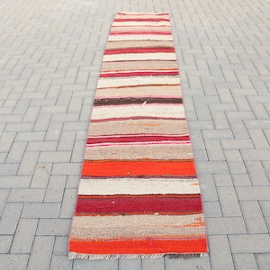 May include: A long, narrow, striped rug with a red, orange, brown, and white color scheme. The rug is made of woven fabric and has a fringed edge.