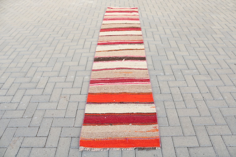 May include: A long, narrow, striped rug with a red, orange, brown, and white color scheme. The rug is made of woven fabric and has a fringed edge.