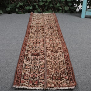 May include: A long, narrow runner rug featuring a complex geometric pattern in shades of rust, brown, beige, and cream. The rug has a repeating symmetrical design throughout its length, finished with short, light-colored fringe at both ends.