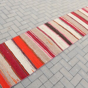 May include: A long, narrow, striped rug with a cream background and red, orange, pink, and brown stripes. The rug has a fringe on both ends.