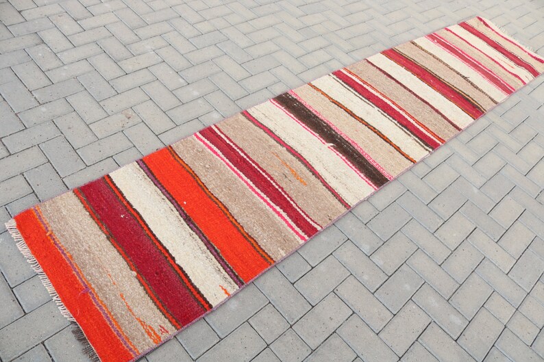 May include: A long, narrow, striped rug with a cream background and red, orange, pink, and brown stripes. The rug has a fringe on both ends.