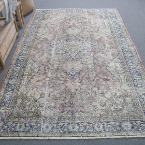 May include: A large, rectangular rug with a faded floral design. The rug's central pattern is in shades of pink, cream, and blue, with a darker blue border. It is placed on a grey surface. The rug has a distressed, vintage appearance.