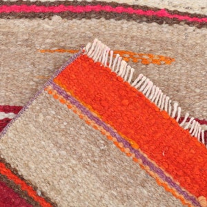 May include: Close-up of a woven rug with a beige background and colorful stripes in shades of red, orange, purple, and brown. The rug has a fringe border.