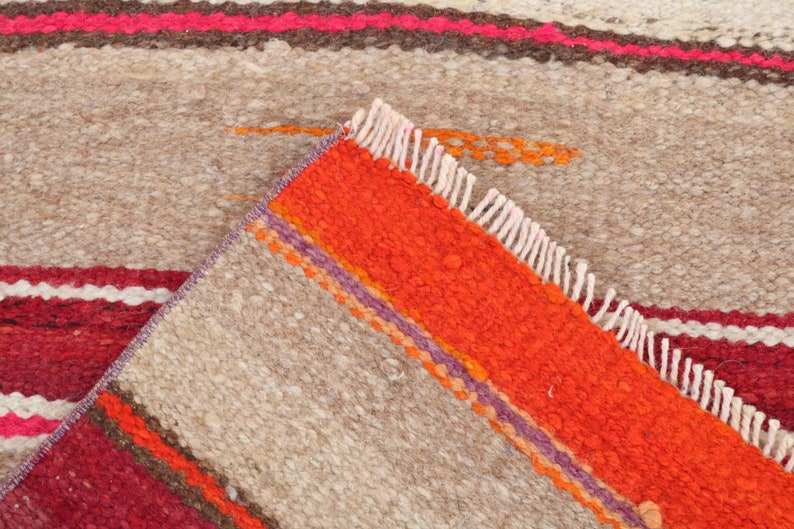May include: Close-up of a woven rug with a beige background and colorful stripes in shades of red, orange, purple, and brown. The rug has a fringe border.