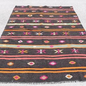 Op de afbeelding: Een zwart en oranje patroonteppich met roze, witte en groene accenten. Het tapijt heeft een franje rand en een geometrisch ontwerp.