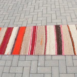 May include: A long, narrow, striped rug with red, orange, brown, and white stripes. The rug is made of a woven material and has a rustic, hand-crafted look.