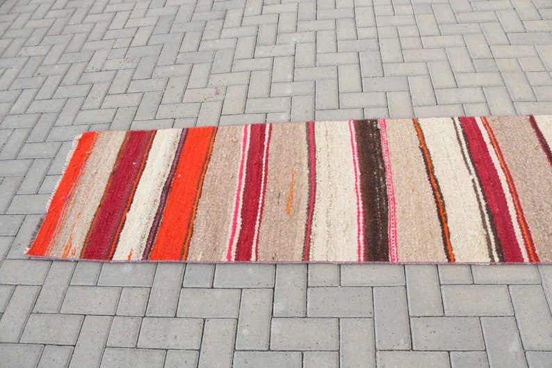 May include: A long, narrow, striped rug with red, orange, brown, and white stripes. The rug is made of a woven material and has a rustic, hand-crafted look.