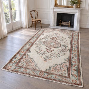 Op de afbeelding: Een rechthoekig vloerkleed met een crèmekleurige basis, een centraal medaillon en ingewikkelde bloemmotieven in tinten terracotta, saliegroen en antraciet. Het kleed heeft een franjerand en een brede, gedetailleerde rand.