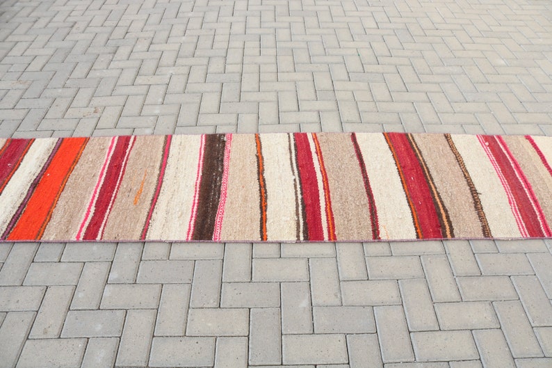 May include: A long, narrow, striped rug with red, orange, brown, and white stripes. The rug is made of a woven material and has a rustic, hand-crafted look.