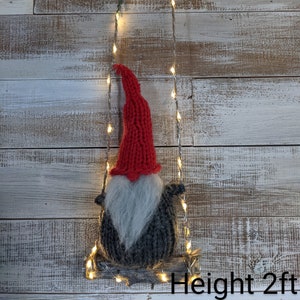 May include: A knitted gnome with a red hat and gray beard is sitting on a wooden swing. The swing is decorated with a string of white lights. The gnome is 2 feet tall.