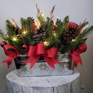 May include: A silver metal planter with a rustic wood base, filled with a Christmas centerpiece. The centerpiece features pine branches, pine cones, red ornaments, and white lights. The planter is decorated with snowflake cutouts and a red satin bow.