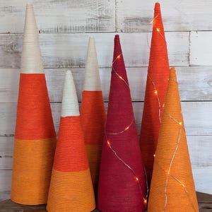 May include: A collection of decorative cone-shaped objects in various sizes and colors. Some cones are orange, white, and yellow, resembling candy corn. Others are solid red or orange, with one adorned with string lights. The cones are arranged on a wooden surface.