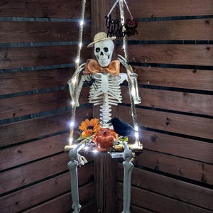 May include: A skeleton figure wearing a straw hat and an orange bow tie sits on a rope swing illuminated by string lights. The swing features a black Happy Halloween sign, a sunflower, a small orange pumpkin, a black crow, and a spider.