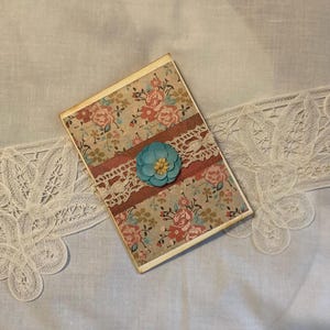May include: A handmade card with a floral pattern and a blue flower with a yellow center. The card is decorated with lace and a red ribbon.