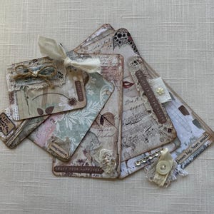 May include: A collection of handmade paper tags with a vintage aesthetic. The tags feature floral and script designs, lace, and embellishments. Some tags have text that reads "BEAUTIFUL", "imagination", and "CRAFT YOUR HAPPINESS".
