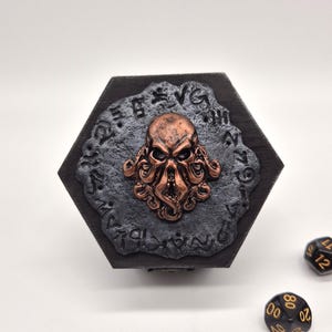 Könnte beinhalten: Schwarze Holzkiste mit einem sechseckigen Deckel, der mit einem kupferfarbenen Krakenkopf mit Tentakeln verziert ist. Die Kiste ist von schwarzen Würfeln mit goldenen Zahlen umgeben.