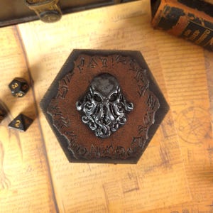Könnte beinhalten: Ein sechseckiger Untersetzer aus braunem Leder mit einem erhabenen Cthulhu-Design aus dunklem Metall. Der Untersetzer hat einen geprägten Schriftzug am Rand. Zwei Würfel sind im Hintergrund zu sehen.