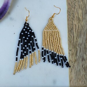 Puede incluir: Un par de pendientes de cuentas con ganchos dorados. Un pendiente presenta un patrón de cuentas negras y blancas con flecos dorados, mientras que el otro es dorado con detalles en negro y blanco. Los pendientes están sobre una superficie blanca y con vetas de madera.