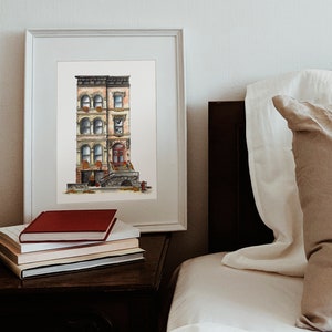 May include: A watercolor painting of a three-story brownstone building with a red door and a stone stoop. The building has windows with white trim and flower boxes. The painting is framed in a white frame and is hanging on a white wall.