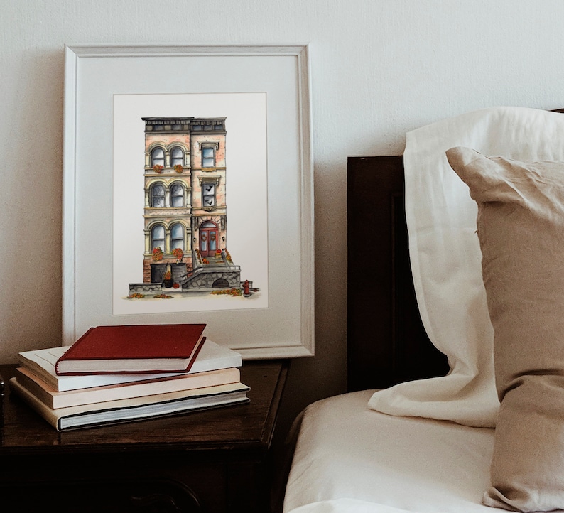 May include: A watercolor painting of a three-story brownstone building with a red door and a stone stoop. The building has windows with white trim and flower boxes. The painting is framed in a white frame and is hanging on a white wall.