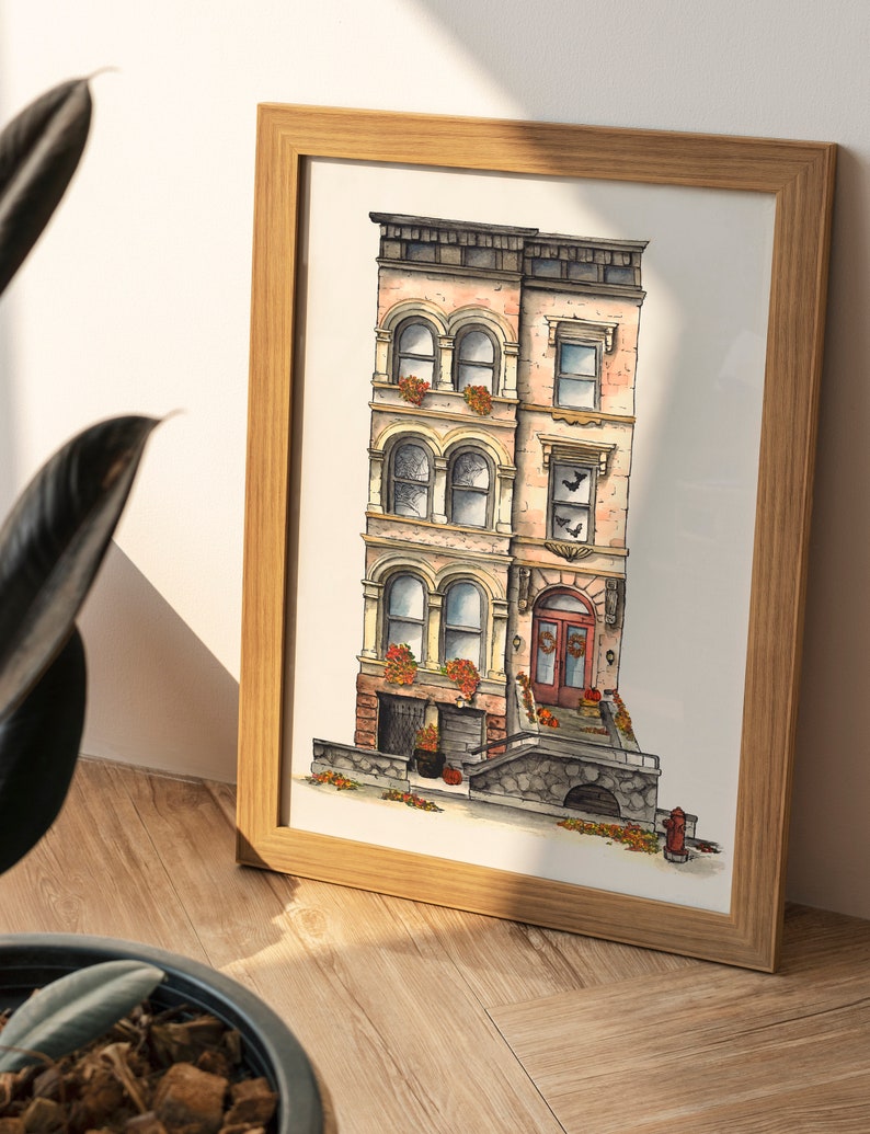 May include: A framed watercolor illustration of a three-story brick building with a brown door and a stone stoop. The building has a pink facade and windows with white trim. There are pumpkins and fall leaves on the stoop.