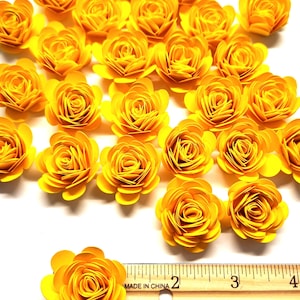 May include: A collection of 25 yellow paper flowers, each measuring approximately 1 inch in diameter. The flowers are arranged on a white surface with a wooden ruler for scale. "Made in China" is printed on the ruler.
