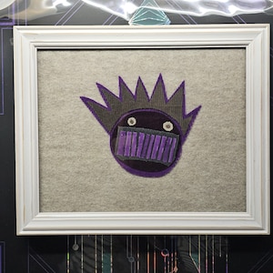 May include: A framed artwork featuring a purple and gray embroidered design of a face with a spiky hairdo and a purple mouth.