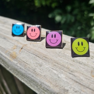 May include: Four square pins with black frames and different colored smiley faces. The pins are arranged in a row on a wooden surface. The colors of the smiley faces are blue, pink, purple, and green.