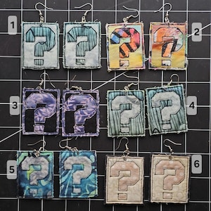 May include: A collection of handmade fabric earrings, each featuring a question mark design. The earrings showcase a variety of colors and patterns, including blue, purple, and orange. The square-shaped earrings are attached to silver-colored ear wires.