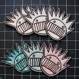 May include: Six embroidered patches featuring a stylized character with a spiky crown and a wide, toothy grin. The top row includes three off-white patches, while the bottom row features one light blue, one grey, and one light pink patterned patch.