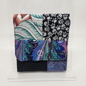 May include: A multi-colored fabric wallet with a patchwork design. The wallet has a black, white, and green floral pattern, a blue and purple marbled pattern, and a black and white geometric pattern. The wallet has a silver metal ring on the front.