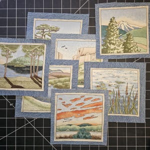 May include: A collection of small, square fabric art pieces featuring landscape scenes. Each piece has a printed image of a natural landscape, framed by a denim border. The scenes include trees, mountains, water, and sunsets.