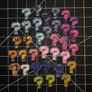May include: A collection of fabric question mark patches in various colors including teal, pink, purple, orange, yellow, and green. Each patch features a stitched border and a unique patterned or marbled fabric design, arranged in a loose cluster.