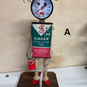 May include: A whimsical sculpture crafted from vintage sewing machine oil can, pressure gauge, and wooden elements. The figure features a red bow, a gauge face, and a red bucket. A measuring tape and red heels complete the design. The oil can reads "SINGER sewing machine OIL".