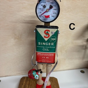 May include: A whimsical, handcrafted sculpture made from repurposed materials. It features a gauge for a head, a Singer sewing machine oil can for a body, and red high-heeled feet. A measuring tape and a small bucket with sequins are included.