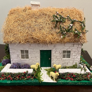 May include: A miniature white cottage with a thatched roof, green door, and two windows. The cottage is surrounded by green grass, red berries, and white stone walls. There are three small white sheep in front of the cottage.