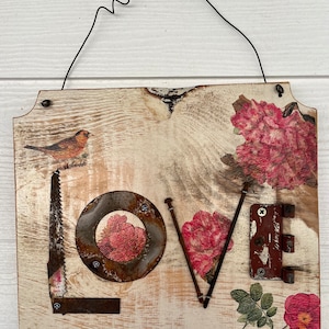 May include: A rustic wooden sign with the word "LOVE" spelled out using various metal objects, including a saw blade, a circular saw blade, nails, and a hinge. The sign is decorated with pink floral patterns and a small bird.